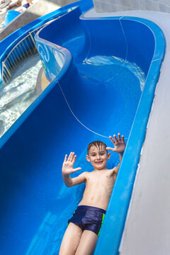 Happy young boy waving hands while sliding down a blue water slide at an indoor water park