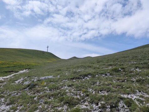 Pendio erboso con rocce affioranti e una grande croce sulla cima, sotto cielo parzialmente nuvoloso