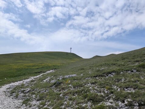 Pendio erboso con rocce affioranti e una grande croce sulla cima, sotto cielo parzialmente nuvoloso