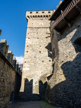 Low-angle view of a historic fortress featuring a tall stone keep tower, fortified battlement walls casting long shadows, an overhanging wooden balcony, and a clear blue sky.