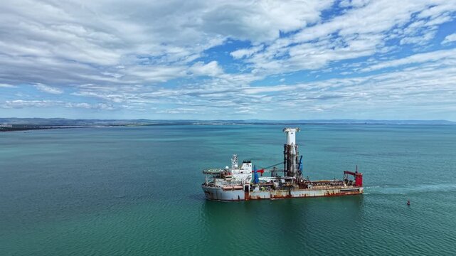 A large industrial dredging vessel works in open waters, moving through calm seas under a wide sky filled with clouds. The vessel is performing dredging activities.