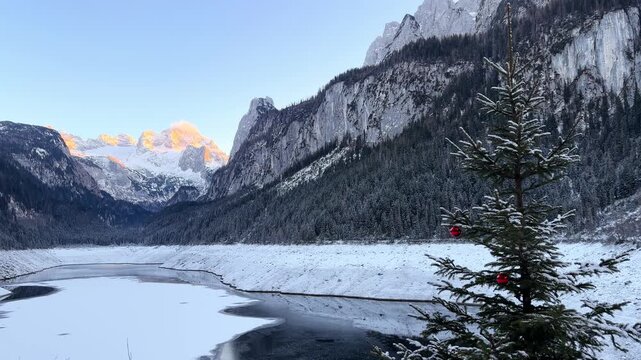 Cinematic sunset over Alpine lake in winter mountains. Winter mountain landscape and Christmas tree with decoration. Alpine peaks illuminated by orange light of setting sun.