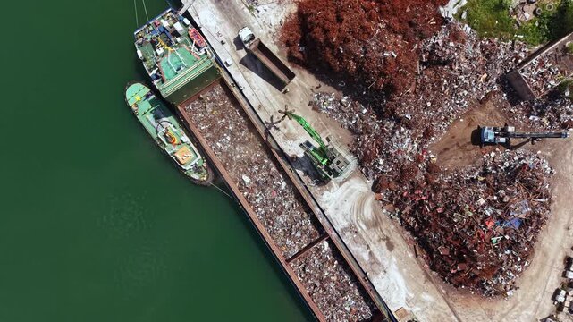 Two boats are docked at a recycling yard. A crane lifts scrap metal from the dock to a pile nearby. Heavy machinery is operating in a busy work environment.