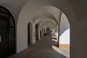 Fototapeta premium Litomysl, Czech Republic - May 1, 2026 - White arched colonnade walkway with stone pavement and dappled sunlight
