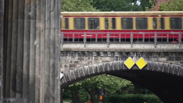 A red and yellow Berlin S-Bahn train crosses a stone bridge above green trees. Motion blur, railings and river architecture create a dynamic city transit view.