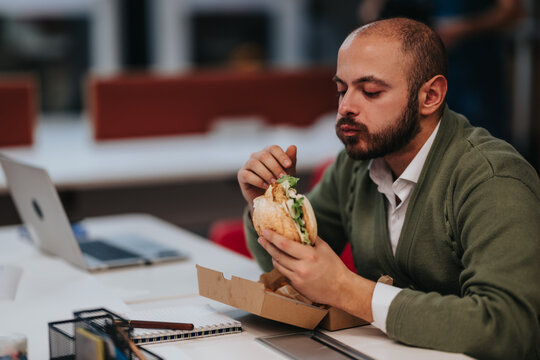 A businessman eats a burger at his office desk during a working lunch. He pauses from work to enjoy a quick meal beside his laptop and notebook