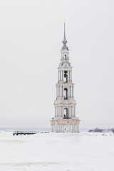 Obraz premium Vertical photo of a tall white bell tower rises alone on a frozen, snow-covered Volga river beneath an overcast winter sky. Landscape with the Flooded Belfry, Kalyazin, Tver Oblast, Russia