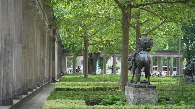 A bronze horse and rider statue stands among trimmed hedges and leafy trees near Berlin's colonnade. The shaded garden blends sculpture, plants and classical architecture.