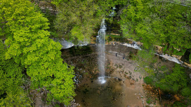 Aerial view of the Herisson waterfalls with water cascading into a rocky pool surrounded by lush green forest and people standing at the base in Menetrux-en-Joux, Bourgogne-Franche-Comte, France.