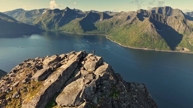 Hiker stands with outstretched arms on steep rocky peak of Segla mountain in Senja, Norway. Deep blue fjord surrounds extreme wild cliffs of nordic island. UHD aerial of summer Senja landscape