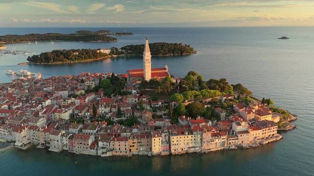 Golden sunset light illuminates terracotta roofs of old town in Rovinj, Croatia. Adriatic sea borders historic church tower on Croatian peninsula. UHD aerial of coastal summer cityscape in Rovinj