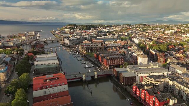 Aerial 4K of colorful historic wooden warehouses lining Nidelva river in Trondheim, Norway. City bridge crosses calm water near traditional waterfront. Nordic skyline of coastal Norwegian cityscape