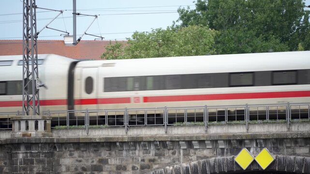 A white ICE train with a red stripe rushes over a stone railway bridge in Berlin. Motion blur, overhead lines and green trees emphasize fast urban transport.