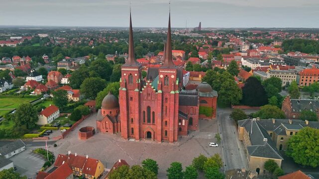 Historic red roofs surround Gothic architecture of Roskilde Cathedral, Denmark in aerial 4K. Medieval brick church dominates urban cityscape. Massive monument stands in central Danish town