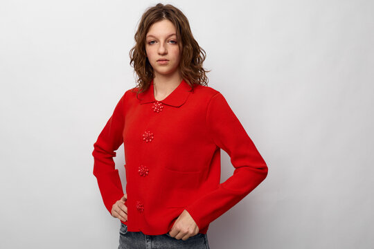 Young woman with curly hair wearing a red sweater and jeans stands against a white background with hands on hips. Casual fashion and confident pose concept.