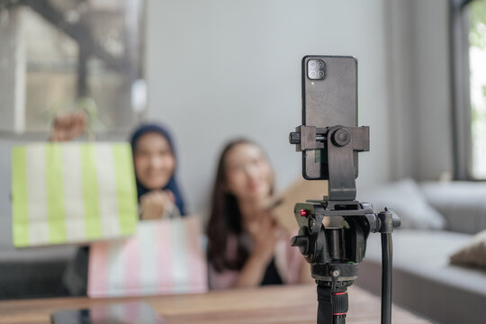 Smartphone mounted on a tripod films a casual review or unboxing scene with two blurred women holding shopping bags in a bright modern interior