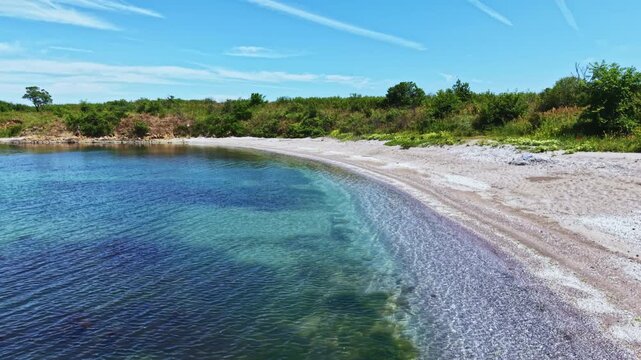 Clear turquoise bay with pebble beach, crystal water lapping curved shoreline under bright blue sky and contrails, sunlit green dunes and sparse vegetation,
