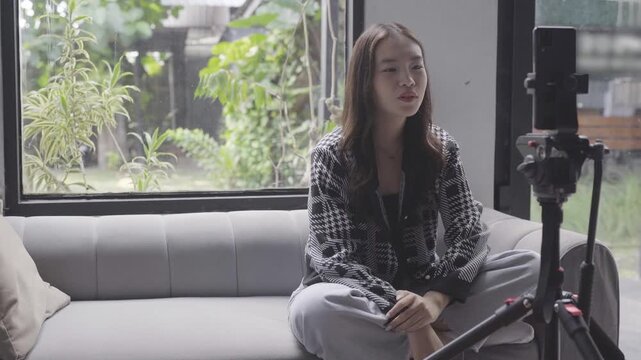 Young asian woman vlogging on sofa, soft natural light through window, indoor plants visible, sitting crosslegged, gesturing