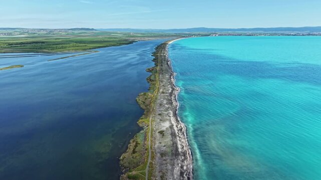 A long stretch of land separates the dark water on one side from the bright blue water on the other side. The view shows clear skies and green land in the background.