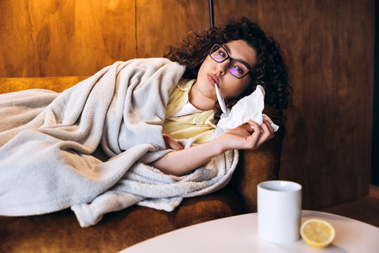 Sick beautiful woman lying under blanket checking temperature cowering with blanket