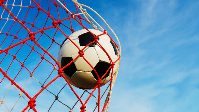 Soccer ball hitting the red and white net scoring a goal during football match with beautiful blue sky background