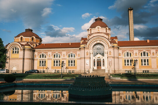 Frontal view of the historic Central Mineral Baths building in Sofia, showcasing its vibrant secessionist facade and stone domes reflected on the fountain pool under a summer sky, Bulgaria