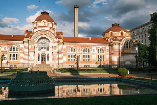 Frontal view of the historic Central Mineral Baths building in Sofia, showcasing its vibrant secessionist facade and stone domes reflected on the fountain pool under a summer sky, Bulgaria
