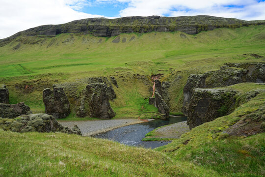 A serene natural landscape featuring a small river flowing through moss-covered rocks and green hills under a partly cloudy sky.,ICELAND