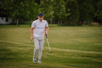 Obraz premium Male golfer in gray pants and white polo shirt walking on green grass course, holding golf club, with trees and a house visible in the background