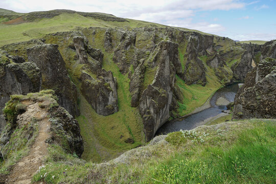 A scenic view of a deep canyon with rugged rock formations, green grassy slopes, and a winding river below.,ICELAND