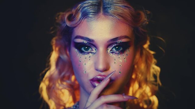 portrait beauty face fashion model posing blonde hair woman gesture of keeping secret silence vibrant make up shiny rhinestones glitter shadows. Adult girl looking at camera eyes art creative make-up