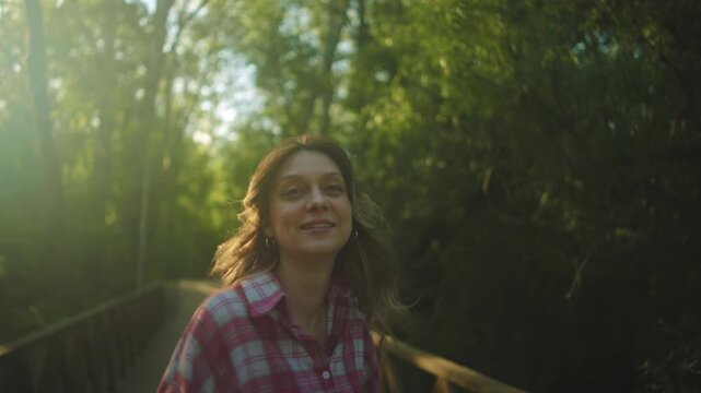 Back view of a young woman standing still on a forest trail surrounded by dense green trees, with warm sunlight flaring from the upper left and creating