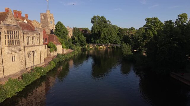 Beautiful historic Gothic church by the river Medway at sunset in Maidstone. Aerial view flying towards pedestrian bridge. Pedestrians walking over the bridge. Lush green trees and old stone buildings