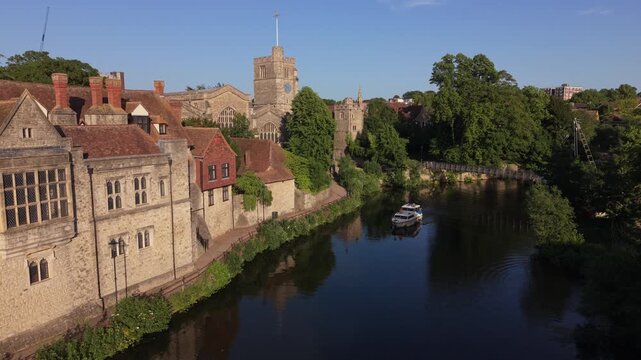 Boat sailing on the river Medway slowly passing by beautiful gothic church in Maidstone at sunset. Aerial view of historic landmark by the river with lush green trees and the bridge for pedestrians.