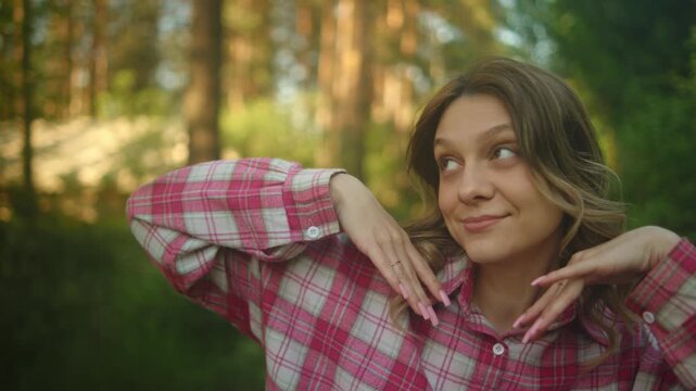 Young woman with wavy hair and a pink plaid shirt stands outdoors in warm natural light, posing with both hands near her face against a soft green woodland