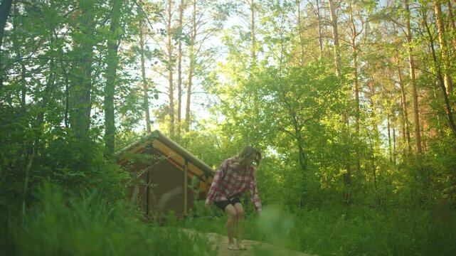 A woman stands on a narrow path in a lush green woodland next to a small wooden cabin, surrounded by tall trees, fresh summer foliage, and soft backlighting