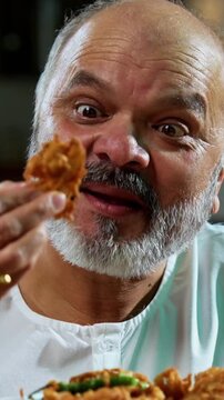 Senior Indian man eating pakoda or pakora with tea on table, Asian foodie old man expressing immense happiness while enjoying tasty onion fritters in modern kitchen setting, joyful retired person