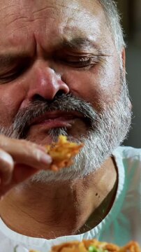 Senior Indian man eating pakoda or pakora with tea on table, Asian foodie old man expressing immense happiness while enjoying tasty onion fritters in modern kitchen setting, joyful retired person