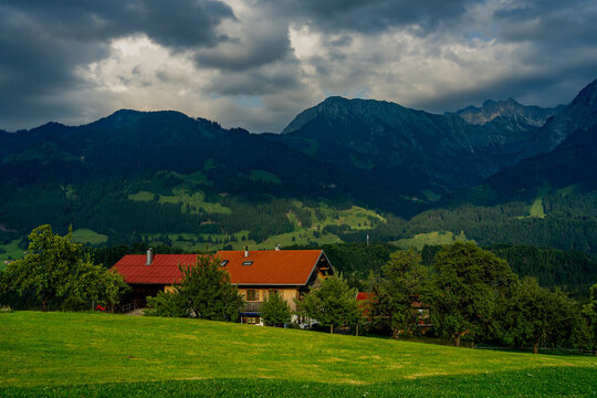 A landscape in the Allg&auml;u region beneath dramatic dark clouds in summer - a traditional house can be seen in the foreground hidden behind trees
