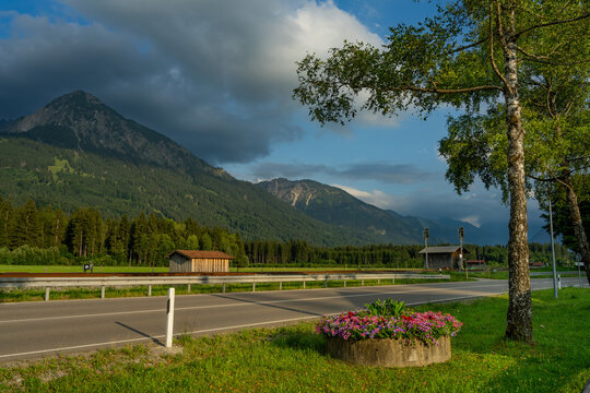 A bowl filled with pink flowers stands in the evening sunlight beside a main street in Langenwang in the Allg&auml;u; mountains can be seen in the background; it is summer