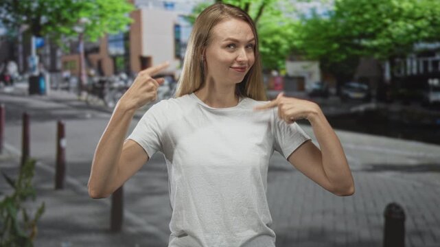 Woman points finger to chest on a city street, smiling and gesturing to herself; confidence self identity.