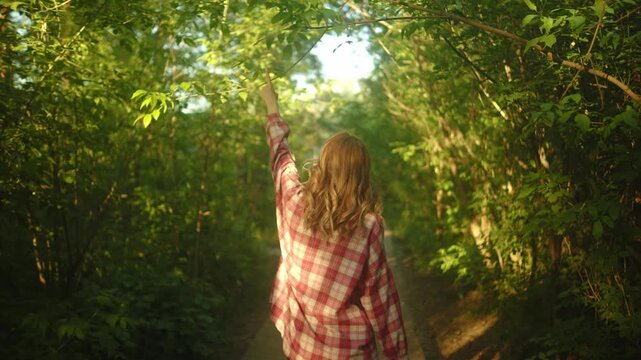 Rear view of a woman walking slowly on a narrow woodland trail surrounded by dense green foliage, soft sunlight, and a calm natural setting