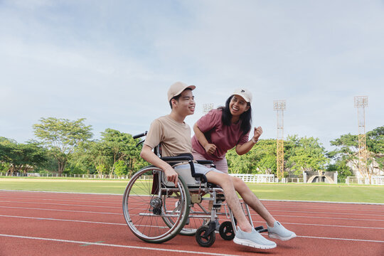 Happy Asian man with disability in wheelchair smiling and talking with supportive mother on running track stadium, diverse family outdoor lifestyle, physical rehabilitation concept.