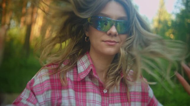 A young woman in a pink plaid shirt stands on a woodland trail, adjusting her sunglasses with both hands. Tall trees, dense green foliage, and warm natural