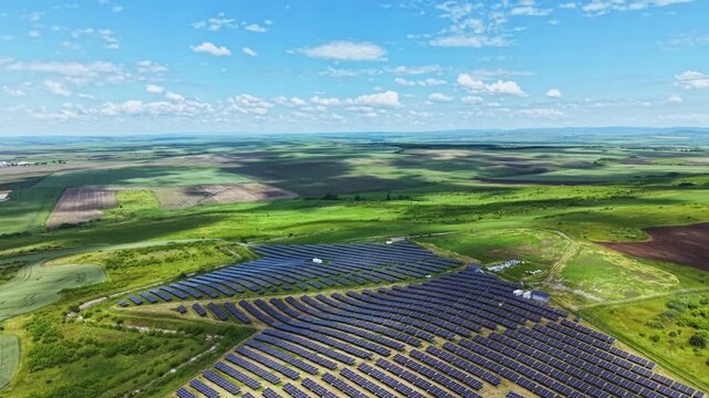 Aerial panorama of solar farm showing rolling green hills, expansive rows of blue panels, distant horizon under scattered clouds drone survey captures scale,