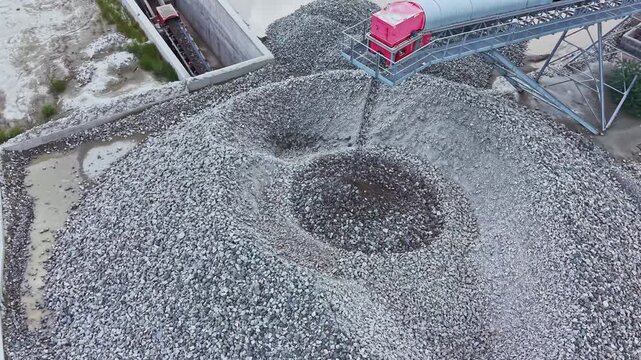 Aerial gravel stockpile receiving conveyor load, red loader depositing stones into central cone, circular depression forming in pile, overhead industrial yard