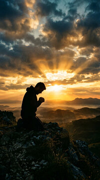 Silhouette of kneeling man in prayer on mountain top at sunset