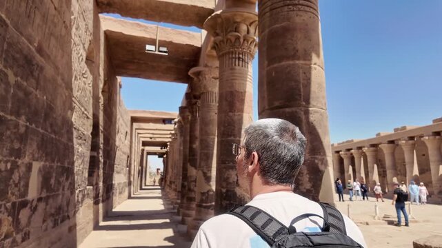 Middle aged tourist man with a backpack walking through the colonnade of the philae temple, admiring ancient egyptian hieroglyphs in aswan