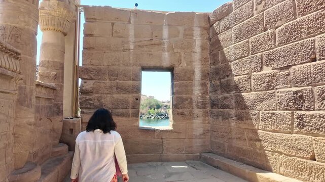 Tourist woman walking inside the ancient Philae Temple, looking out a window at the Nile River. A famous historical landmark in Aswan, Egypt