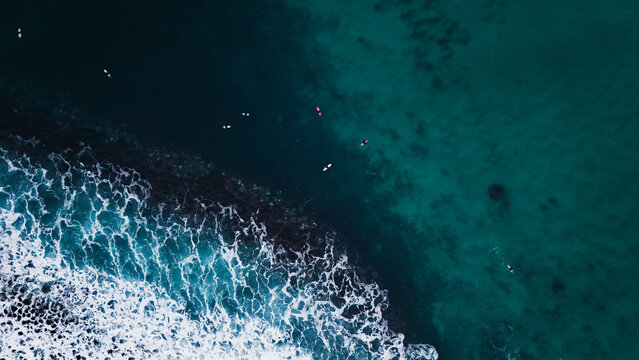Aerial view of surfers waiting for waves in deep turquoise water near a white breaking wave in Unstad, Nordland, Norway.